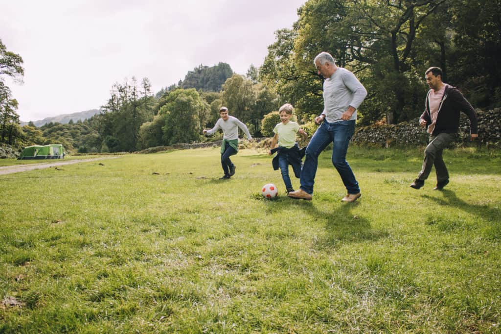 Three generation family are playing football together in a field. There are two boys, their father and their grandfather.
